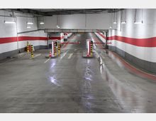 Ein beleuchtetes Parkhaus mit Betonboden und rot-weißen Markierungen an den Wänden. Im Vordergrund befinden sich Schranken und Sicherheitspfosten, dahinter eine ansteigende Rampe mit Fahrbahnmarkierungen.