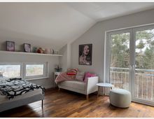 Bright attic bedroom with a sloped ceiling, wooden floors, a low bed beneath small horizontal windows and a long wall shelf. A white sofa with pillows sits by a large glass door to a balcony overlooking trees, with a small side table and round ottoman nearby.