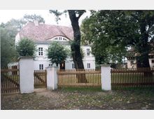 Ein zweistöckiges historisches Gebäude mit weißer Fassade und rotem Ziegeldach, umgeben von Bäumen. Im Vordergrund befindet sich ein Holzzaun mit Tor, dahinter eine Wiese mit großen Bäumen.