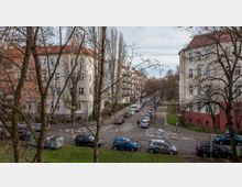 Wohnstraße in einem städtischen Viertel mit mehrstöckigen Wohnhäusern im Stil des frühen 20. Jahrhunderts, hellen Fassaden und roten Dächern. Die Straße ist von geparkten Autos gesäumt, und Bäume ohne Laub stehen im Vordergrund.