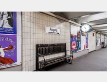 U-Bahn-Station mit gefliesten Wänden, darauf zahlreiche Plakate. Eine schwarze Bank steht unter einem Schild mit der Aufschrift „Ausgang“, daneben hängt eine runde Uhr.