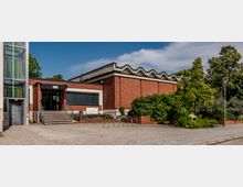 Backsteinbau mit modernem Flachdach und gezacktem Dachrand, daneben ein verglaster Aufzugsschacht. Vor dem Gebäude befinden sich eine kleine Treppe, Fahrradständer und Grünflächen mit Bäumen und Sträuchern.