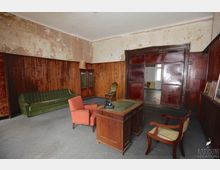 Ein einfacher, altmodischer Raum mit Holzvertäfelungen und abblätternder Wandfarbe. Die Einrichtung umfasst ein grünes Sofa, einen roten Sessel, einen Holztisch mit Telefon und eine Glasvitrine; im Hintergrund ist eine geöffnete Schiebetür aus dunklem Holz zu sehen, die in einen angrenzenden, unmöblierten Raum führt.