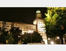 Nachtszene eines großen historischen Gebäudes mit hell erleuchteten Fenstern und einem beleuchteten Turm an der Ecke. Im Vordergrund sind Bäume zu sehen, die teilweise von Straßenlaternen angestrahlt werden.