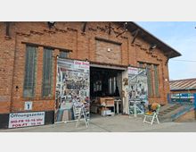 Backsteinfassade einer ehemaligen Industriehalle mit hohen Glasbausteinfenstern und großer, teils geöffneter Toröffnung, an der Werbebanner für einen Trödelmarkt hängen. Vor dem Eingang stehen Aufsteller und einzelne Möbelstücke; rechts führt eine Rampe mit blauem Geländer entlang des Gebäudes.