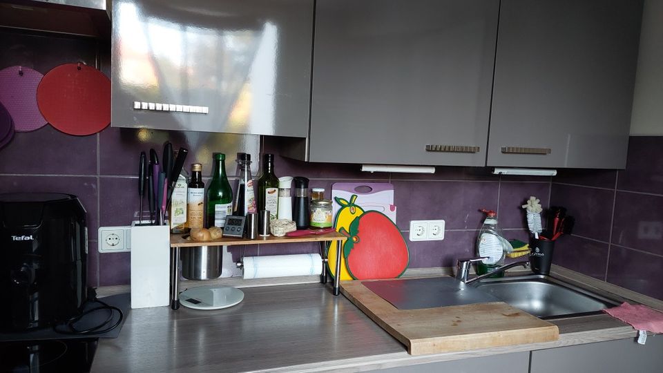 Kompakte Küchenzeile mit grauen Hängeschränken, lila Fliesenrückwand und Arbeitsplatte mit kleinem Regal voller Flaschen und Küchenutensilien. Rechts befindet sich eine Edelstahlspüle mit Schneidebrett und Abtropffläche, links ein schwarzes Küchengerät auf der Arbeitsfläche.