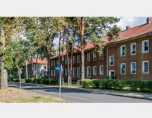 Straßenansicht mit zweigeschossigen, braun verputzten Wohngebäuden und roten Ziegeldächern, umgeben von hohen Kiefern und niedrigen Hecken. Ein Straßenschild zeigt den Namen „Mozartstr.“, und die Umgebung ist von Bäumen und Grünflächen geprägt.