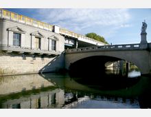 Steinerne Brücke über einen Fluss mit einem angrenzenden Gebäude in klassischer Architektur und einer Statue auf dem Brückengeländer; im Hintergrund Bäume und ein klarer Himmel.