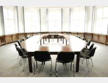 Großer Besprechungsraum mit U-förmigem Holztisch und gepolsterten Stühlen, helle Wände mit großen Fenstern und hölzernen Heizungsverkleidungen entlang der Wände.