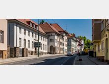 Straßenansicht einer Stadt mit mehrstöckigen Wohn- und Geschäftshäusern, teilweise verputzt oder mit Ziegeldecken. Rechts führt eine einspurige Straße mit Gehwegen entlang, auf der Autos und Verkehrsschilder zu sehen sind.