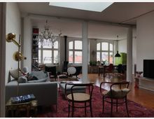 Wohnbereich eines Lofts mit Holzfußboden, einem großen roten Teppich und hohen weißen Wänden. Es gibt einen grauen Sofa-Bereich, eine offene Ess- und Arbeitszone mit grünen und bunten Stühlelementen, große Fenster mit Blick auf Dächer und eine zentrale Kristall-Kronleuchterlampe.