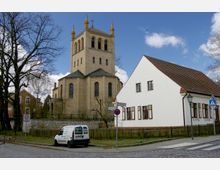 Historische Backsteinkirche mit einem Glockenturm und hohen Fenstern, umgeben von einem Zaun und Bäumen, in einer ländlichen Ortschaft. Rechts im Vordergrund ein weißes Gebäude mit roten Dachziegeln und Holzfenstern sowie ein gepflasterter Gehweg und ein Verkehrsschild.