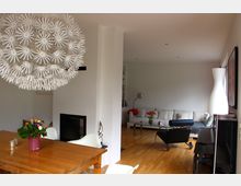 Wohnzimmer in einem modernen Haus mit Holzboden und klaren, weißen Wänden. Im Vordergrund ein Esstisch mit Blumenstrauß und auffälliger Lampenschirm in Blütenform; im Hintergrund eine Sitzecke mit weißem Sofa, Couchtisch und Design-Sessel.