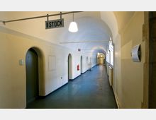 Langer, schlichter Flur in einem historischen Gebäude mit gewölbter Decke, mehreren Türen mit Rundbögen auf der linken Seite und Fensterfronten auf der rechten Seite. An der Decke hängt ein Schild mit der Aufschrift „II STOCK“.