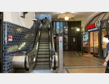 Eine Rolltreppe in einer Bahnhofshalle, flankiert von dunkelblau gefliesten Wänden links und einem Bäckereigeschäft mit Glasfront rechts. Im Hintergrund sind Eingangstüren und große Fenster mit modernem Design sichtbar.