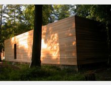 Rechteckiges, modernes Gebäude mit einer Fassade aus horizontalen Holzbrettern, umgeben von hohen Bäumen in einem bewaldeten Bereich. Die Sonnenstrahlen werfen Schatten der Bäume auf die Holzfassade.