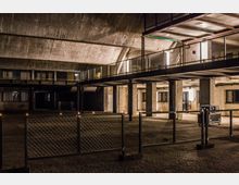 Industriehalle mit grobem Betongewölbe, Stahlträgern und Backsteinwänden. Im Vordergrund befindet sich ein Gitterzaun, dahinter ein zweistöckiger Bereich mit Geländern und Arbeitsräumen.