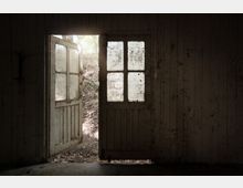 Blick aus dem Inneren eines dunklen, abgenutzten Holzgebäudes durch eine geöffnete Doppeltür mit Glasfenstern auf einen erdigen Hang im Freien, durch den Tageslicht einfällt. Die Holzwände und die Türen wirken alt und verwittert.