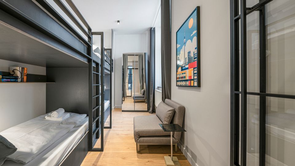 Schmales, modern eingerichtetes Schlafzimmer mit übereinander angeordneten Metall-Etagenbetten, hellem Holzboden und grauen Vorhängen an hohen Fenstern. An der rechten Wand stehen ein kleines graues Sofa mit Beistelltisch und ein farbiges Bild; Glastrennwände mit schwarzen Rahmen führen zu einem Flur im Hintergrund.