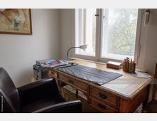 Arbeitszimmer mit einem rustikalen Holztisch vor einem großen Fenster, darauf eine Schreibtischunterlage, eine Lampe, Schreibutensilien und ein Drucker. Im Vordergrund ein Ledersessel, an der Wand ein abstraktes Gemälde.