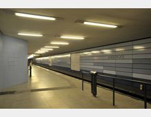 U-Bahn-Station mit glatten Wänden aus gestreiften blauen und weißen Paneelen, Deckenbeleuchtung und Fliesenboden. An der Wand ist der Schriftzug "Bundesplatz" zu sehen; im Hintergrund befinden sich ein Bahnsteig und eine Anzeigetafel.
