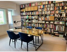 Ein Arbeitszimmer mit einem großen, goldfarbenen Tisch und vier blauen Stühlen. An der Rückwand befindet sich ein deckenhohes Regal, gefüllt mit Büchern und Aktenordnern. Ein Fenster mit weißem Rollo und eine Flipchart stehen auf der linken Seite, daneben ist ein Mikrofon mit Studioausrüstung sichtbar.