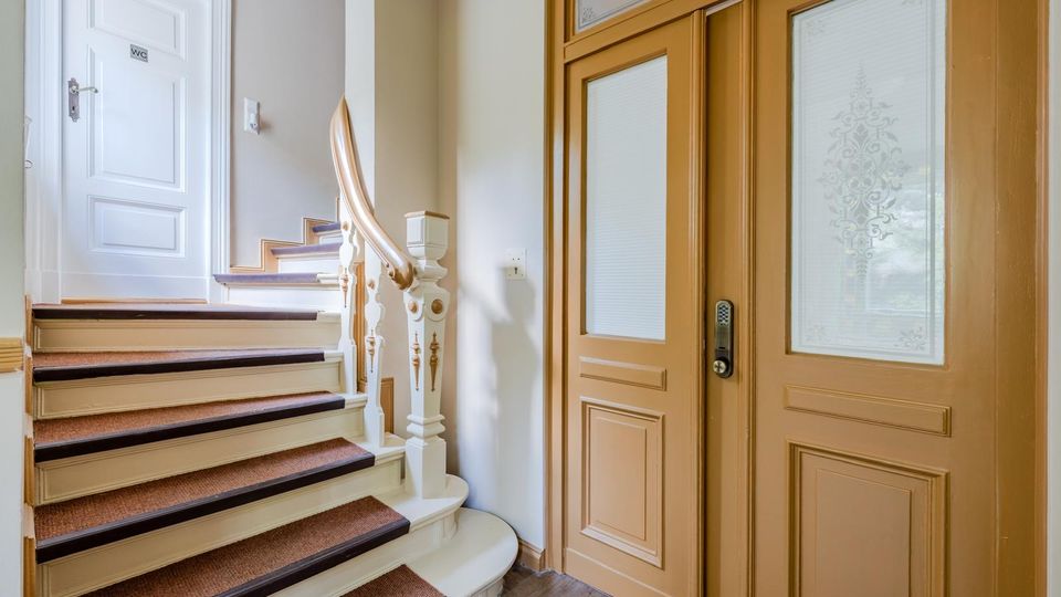 Innenbereich eines Treppenhauses mit gebogener Holztreppe, Teppichläufer und dekorativem Geländer. Rechts stehen große beige Doppeltüren mit Glasfeldern und Ornamentmuster, links eine weiße Tür mit WC-Schild.