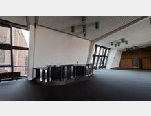Großer, leerer Innenraum mit dunklem Boden, einer zentralen Theke aus Metall und großen Fensterfronten, die den Blick auf ein Backsteingebäude freigeben. Die Decke ist mit rasterförmigen Paneelen und hängenden Leuchten ausgestattet, am Ende des Raums befindet sich eine holzverkleidete Wand mit Durchgang.