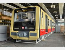 Ein Zug der Berliner S-Bahn (Baureihe 483/484) steht in einer Werkshalle. Der gelb-rote Zug mit der Linienanzeige "S41 Ring" ist auf einem Gestell montiert, umgeben von Regalen und Werkstattinventar.