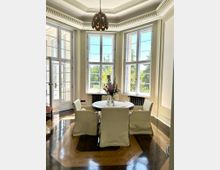 Heller Speiseraum in einem Altbau mit hohen Decken, Stuck und zentraler Hängelampe sowie großen Sprossenfenstern in einem Erker. In der Mitte steht ein runder Tisch mit vier Stühlen auf einem Holzparkettboden; seitlich führt eine Glastür nach außen.