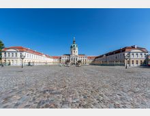 Barockes Schloss mit hellem Fassadenputz und rotem Dach, flankiert von zwei symmetrischen Seitenflügeln. Im Vordergrund ein gepflasterter Hof und schmiedeeiserne Gitter, zentral ein Turm mit grüner Kuppel und vergoldeter Statue auf der Spitze.