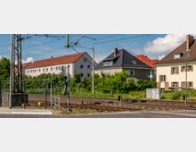 Wohngebiet mit Bahnschienen im Vordergrund, umgeben von Einfamilienhäusern mit roten und dunklen Dächern sowie grüner Vegetation im Hintergrund. Über den Schienen verlaufen Stromleitungen, die an Masten befestigt sind.