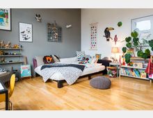 Kinderzimmer mit einem Holzboden, einem weißen Bett mit dekorativen Kissen und Decken sowie einer grauen und weißen Wand. An der Wand hängen Bilder und Tierdekorationen, daneben stehen ein Regal mit Büchern und Spielzeug sowie eine Tischlampe.
