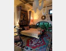 Schlafzimmer mit orientalischem Dekor, ausgestattet mit einem Metallbett, traditionellen gemusterten Teppichen und farbenfrohen Textilien. Die Wände sind mit einem handgefertigten Wandteppich und einem runden Spiegel verziert, während die helle Beleuchtung eine gemütliche Atmosphäre schafft.