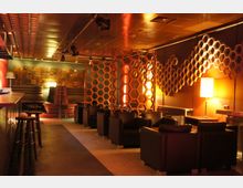 Eine moderne Lounge mit dimmer Beleuchtung, schwarzen Ledersesseln und halbhohen Waben-Trennwänden aus Holz. An den Wänden befinden sich geometrische Holzverzierungen, und die Einrichtung ist in warmen Orangetönen gehalten.