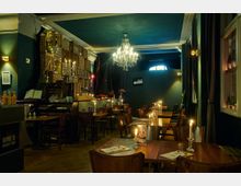 Gemütliche Wein- und Whiskybar mit hölzernen Tischen und Stühlen, dekoriert mit Kerzenhaltern und Kerzen. Der Raum hat dunkelgrüne Wände, einen Kronleuchter, Holzkisten an der Wand und eine warme, indirekte Beleuchtung.