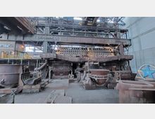 Innenansicht einer stillgelegten Industrieanlage mit einem massiven Stahlgerüst, rostigen Maschinen und Trichtern. Im Hintergrund sind metallische Strukturen, Rohre und ein Kran innerhalb einer großen Halle erkennbar.