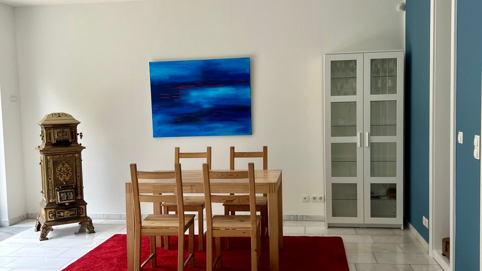 Helles Esszimmer mit weiß gefliesten Bodenplatten, einem Holztisch mit sechs Stühlen auf einem roten Teppich und einem großen blau abstrakten Wandbild. Rechts steht ein weißer Glasvitrinenschrank neben einer blau akzentuierten Wand, links ein dekorativer antiker Ofen.