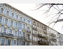 Mehrstöckige Gebäude im Stil des Historismus, mit dekorativen Fassaden, großen Fenstern und kleinen Balkonen; die Fassade eines Gebäudes ist in Blau und Weiß gestrichen, während die danebenliegenden eine bräunliche oder helle Putzfassade haben. Im Vordergrund stehen kahle Bäume.