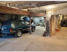 Ein einfacher Lagerraum mit rohen Betonwänden, einer freiliegenden Metallstütze und sichtbaren Kabeln an der Wand. Im Vordergrund steht ein dunkelgrünes Geländewagen, im Hintergrund sind Abdeckplanen, Gerätschaften und ein großer schwarzer Tank sichtbar.