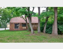 Backsteinbau mit roten Dachziegeln und großen Fenstern, umgeben von mehreren hohen Bäumen und einer gepflegten Rasenfläche. Im Hintergrund dichter grüner Buschbewuchs und ein gepflasterter Weg.