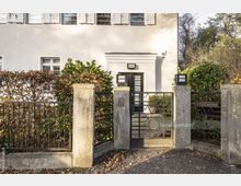 Weiß verputztes Wohnhaus mit schwarzen Fensterläden und einer schwarzen Eingangstür. Vor dem Gebäude befindet sich ein Metallzaun mit Pfeilern aus Beton und einem kleinen Gartentor, das von Pflanzen und Hecken umgeben ist.