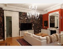 Wohnzimmer mit hellen weißen Möbeln, einer weißen Zierdecke und Holzboden. An der Wand befinden sich dunkle Tapeten mit floralen Mustern, kombiniert mit einer roten Akzentwand und einem Kamin mit weißem Rahmen; ein Kronleuchter hängt zentral von der Decke.