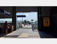 Bahnhofsausgang mit einem überdachten Steg, der ins Freie führt. Menschen spazieren auf dem Steg, hinter dem moderne Gebäude und ein bewachsener Bereich sichtbar sind; Wegweiser und Werbeplakate rahmen den Ausgang.