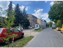 Straßenansicht in einem Wohngebiet mit geparkten Autos auf beiden Seiten, mehrstöckigen Altbauhäusern auf der rechten Seite und großen Bäumen auf der linken Seite. Der Himmel ist teilweise bewölkt, und im Hintergrund sind weitere Grünflächen sichtbar.