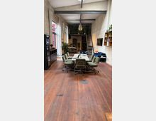 Großer, offener Raum mit Industrieloft-Charakter, ausgestattet mit einem Konferenztisch und mehreren grünen Bürostühlen auf dunklem Holzboden. Im Hintergrund befinden sich eine schmale Metalltreppe, Regale mit Pflanzen und Dekoration sowie hohe Fenster mit weißen Vorhängen.