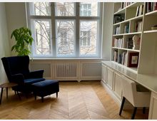 Wohnzimmer oder Leseecke mit einem großen Sprossenfenster, das Tageslicht in den Raum lässt. Links ein schwarzer Sessel mit Fußhocker, daneben eine grüne Zimmerpflanze, rechts ein weißes Regal mit Büchern und Dekorationen sowie ein integrierter Schreibtisch mit einem weißen Stuhl. Der Boden ist aus hellem Holz in Fischgrätmuster.