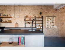 Moderne Küche mit freiliegender Ziegelwand, einem dunklen Inselarbeitsbereich mit Spüle und offenen Regalen mit Geschirr und Dekorationsgegenständen. An der Wand hängt ein Poster mit Pilzillustrationen.