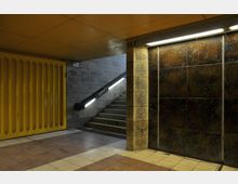 Unterirdischer Durchgang mit grauen Steinwänden, gelbem Metallrolltor auf der linken Seite, einer beleuchteten Treppe in der Mitte und schwarzen Wandfliesen rechts. Der Boden besteht aus beigen und braunen Fliesen.