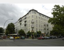 Eckgebäude eines Wohn- und Geschäftshauses mit heller Fassade, mehreren Balkonen und Geschäften im Erdgeschoss, umgeben von geparkten Autos, einem Fahrradfahrer und einer vorbeifahrenden gelben Straßenbahn; Bäume säumen die Straßenseite.